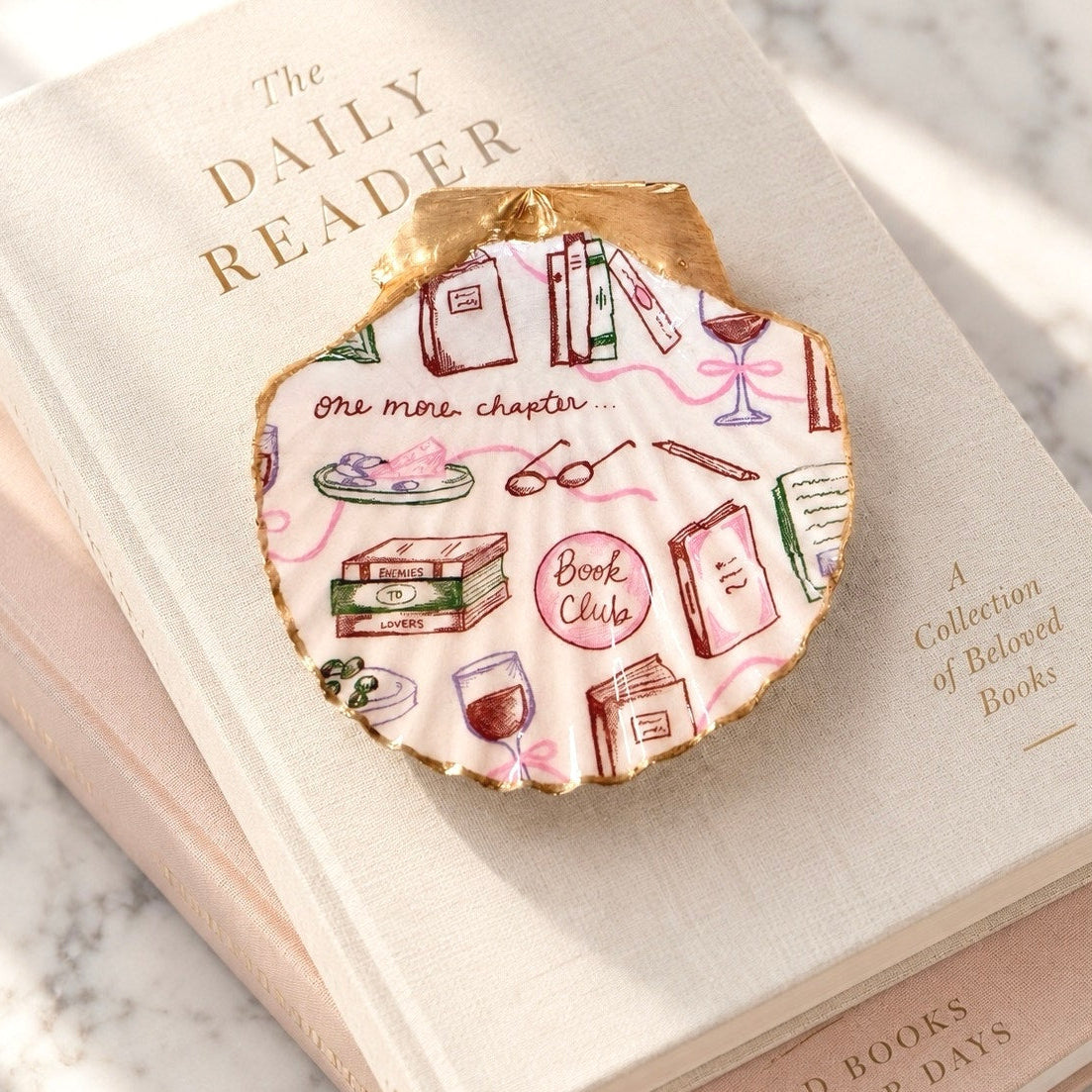 A scalloped-edge paper bookmark with a gold border and a pink-and-white illustrated pattern featuring books, glasses, and the words 'Book Club' and 'One mini-chapter.